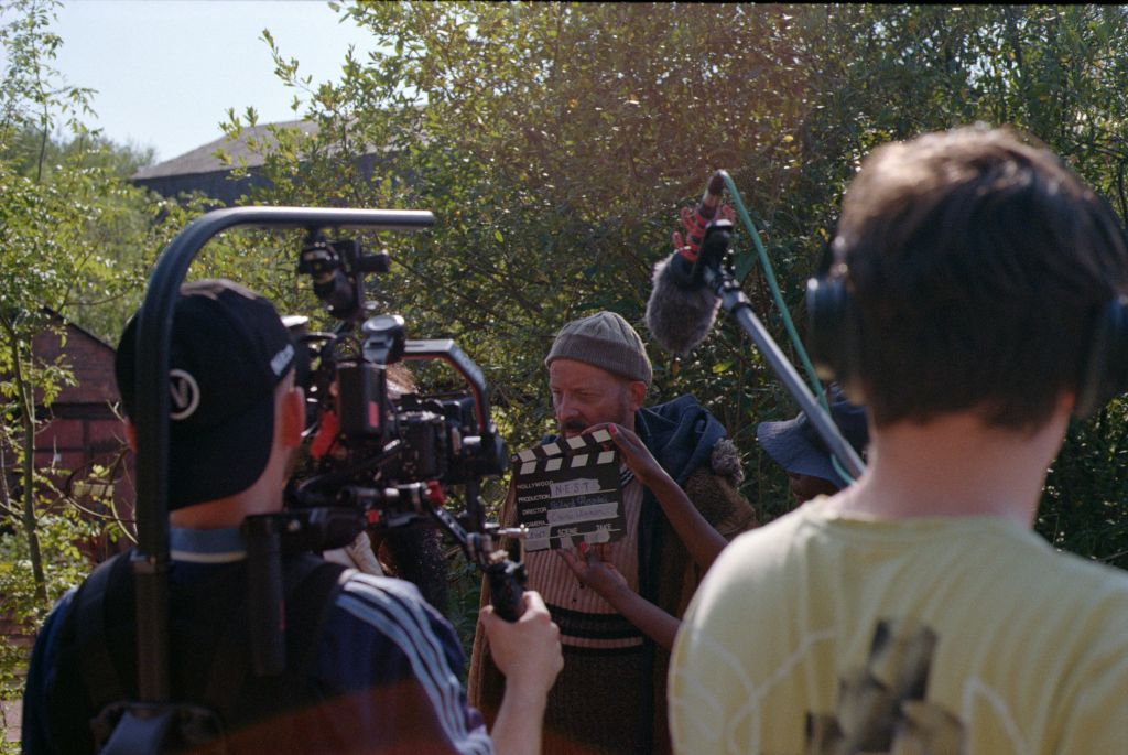 A close up of film crew gathered around an actor, one holds a clapper board, the rest hold cameras and boom microphones.