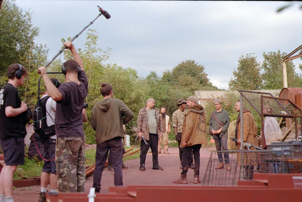 A film crew prepares to capture a scene outdoors on a cloudy day, with trees in the background. A boom operator holds up a microphone on a boom pole while others look on.