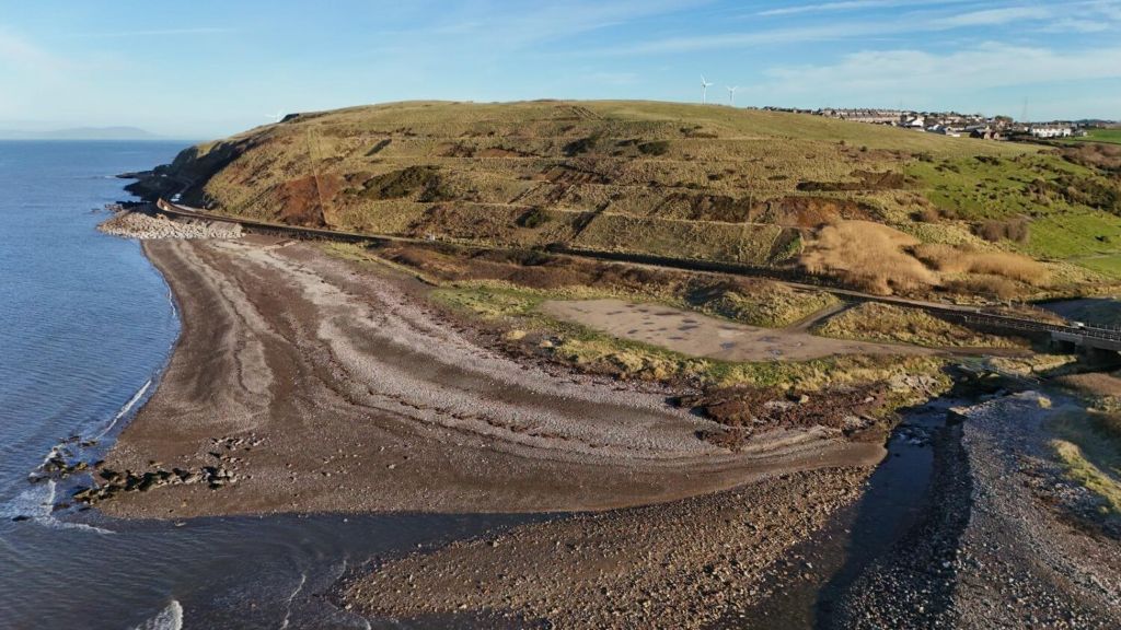 An aerial view of a beach in Flimby with a cliffside, showing the coastline and surrounding landscape on a sunny day