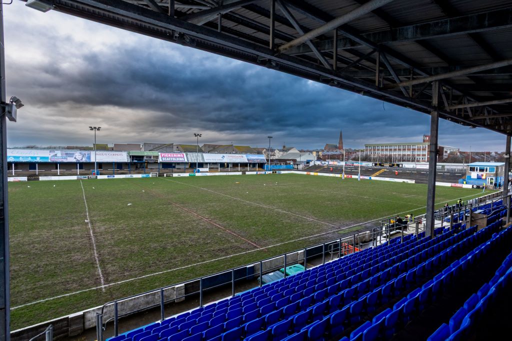 An empty rugby stadium with blue seats on a cloudy day, showing a green field with white lines.